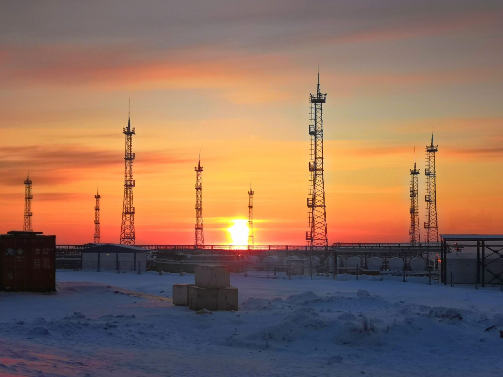 Industrial utility poles silhouetted against a vibrant sunset in a snowy landscape.